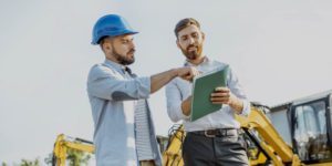 Man in hard hat review documents with another man in front of construction equipment.