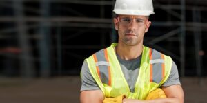 A man standing on a jobsite looks into the camera, wearing a hard hat, safety glasses, a safety vest and work gloves