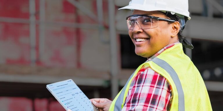 Woman in safety vest and hard hat smiles while holding a tablet.